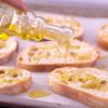 Bật lò trước ở 400 độ C. Bánh mì bạn mua loại baguette, sau đó cắt thành miếng nhỏ dày khoảng 2cm nhé. Sử dụng một cái khay có lót sẵn giấy nến, xếp bánh mì lên. Rưới dầu olive khắp 2 mặt bánh mì.
